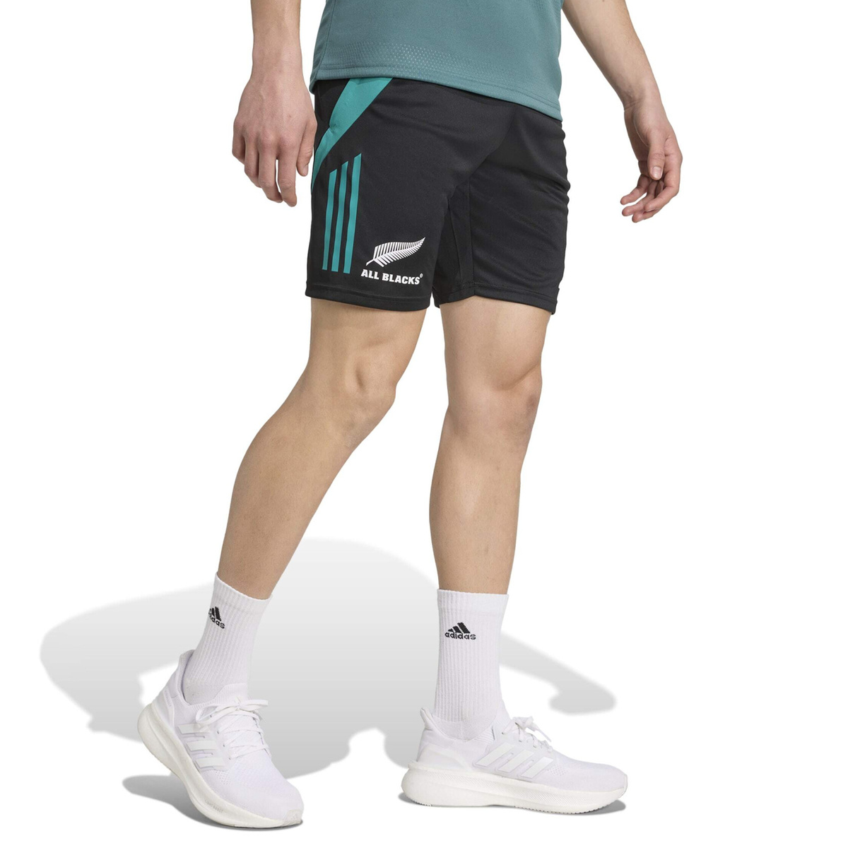 Sporting the adidas All Blacks 25/26 Training Gym Short in black with teal accents, a matching teal shirt, white adidas socks, and sneakers, this person highlights New Zealand rugby style against a white backdrop.