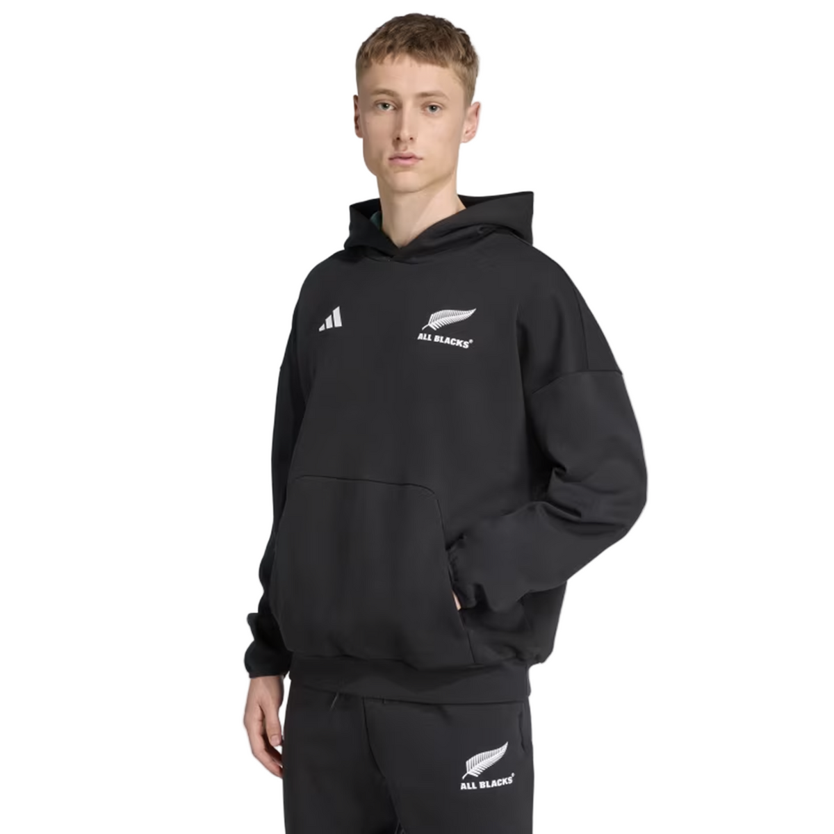 A person wearing the adidas All Blacks 25/26 Presentation Hoodie, featuring the team's logo, along with matching pants stands against a plain white background.