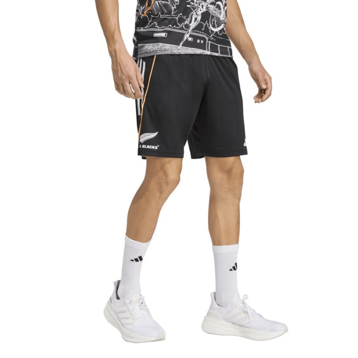 A person wearing All Blacks 26/27 Marvel Performance Gym Shorts by adidas with a white logo, white socks, white sneakers, and a patterned shirt stands against a plain background.