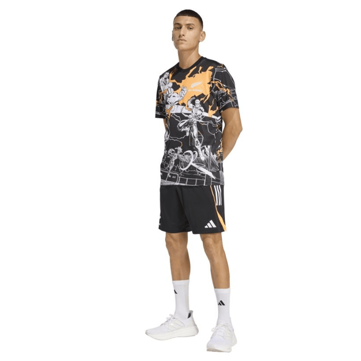 A man faces forward wearing a black graphic tee, adidas All Blacks 26/27 Marvel Performance Gym Shorts with orange accents, white crew socks, and white sneakers.
