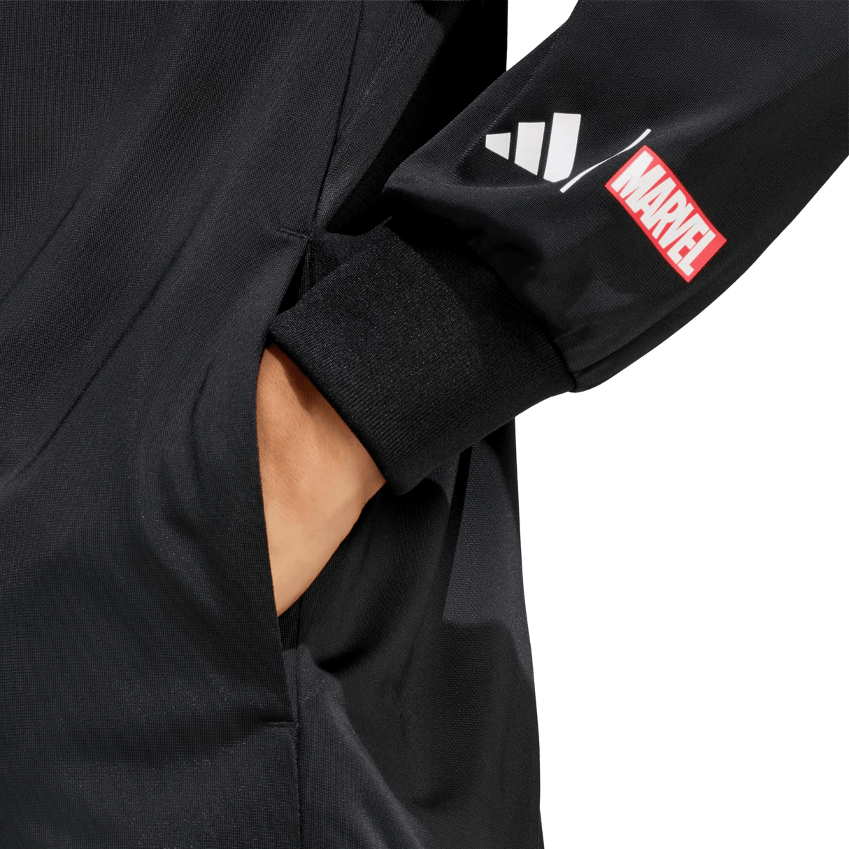 A hand is in the pocket of black pants. The black sleeve, from the adidas All Blacks 26/27 Marvel Track Top, features adidas stripes and a red Marvel logo patch for a bold, Marvel-inspired sportswear style.