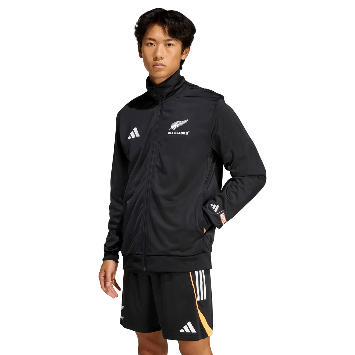A person in the adidas All Blacks 26/27 Marvel Track Top and black shorts with white and gold stripes stands against a plain light background.