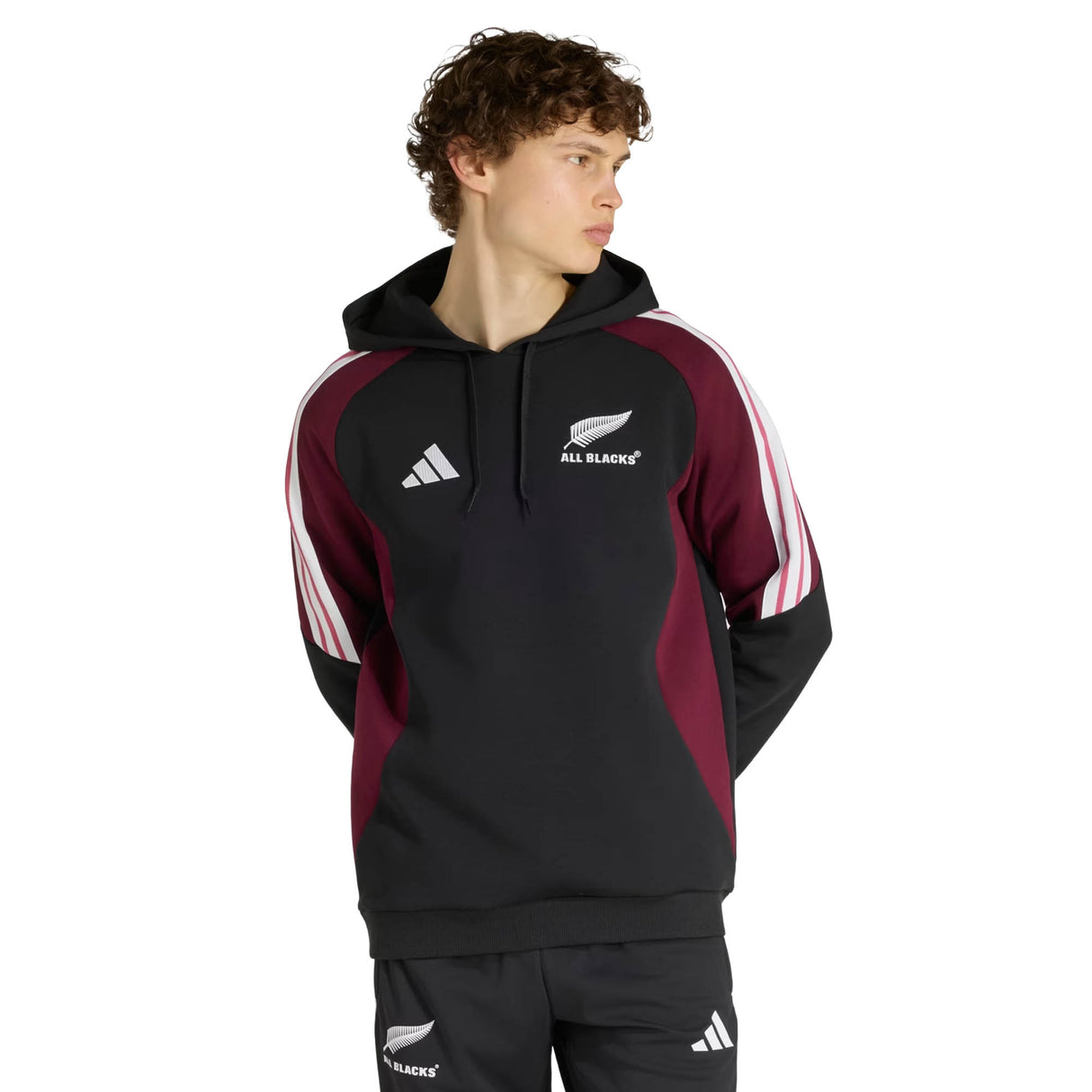 Sporting the adidas All Blacks 27/28 Hoodie in black and maroon, featuring signature white sleeve stripes, a person looks off to the side against a simple backdrop—capturing the timeless spirit of New Zealand rugby.