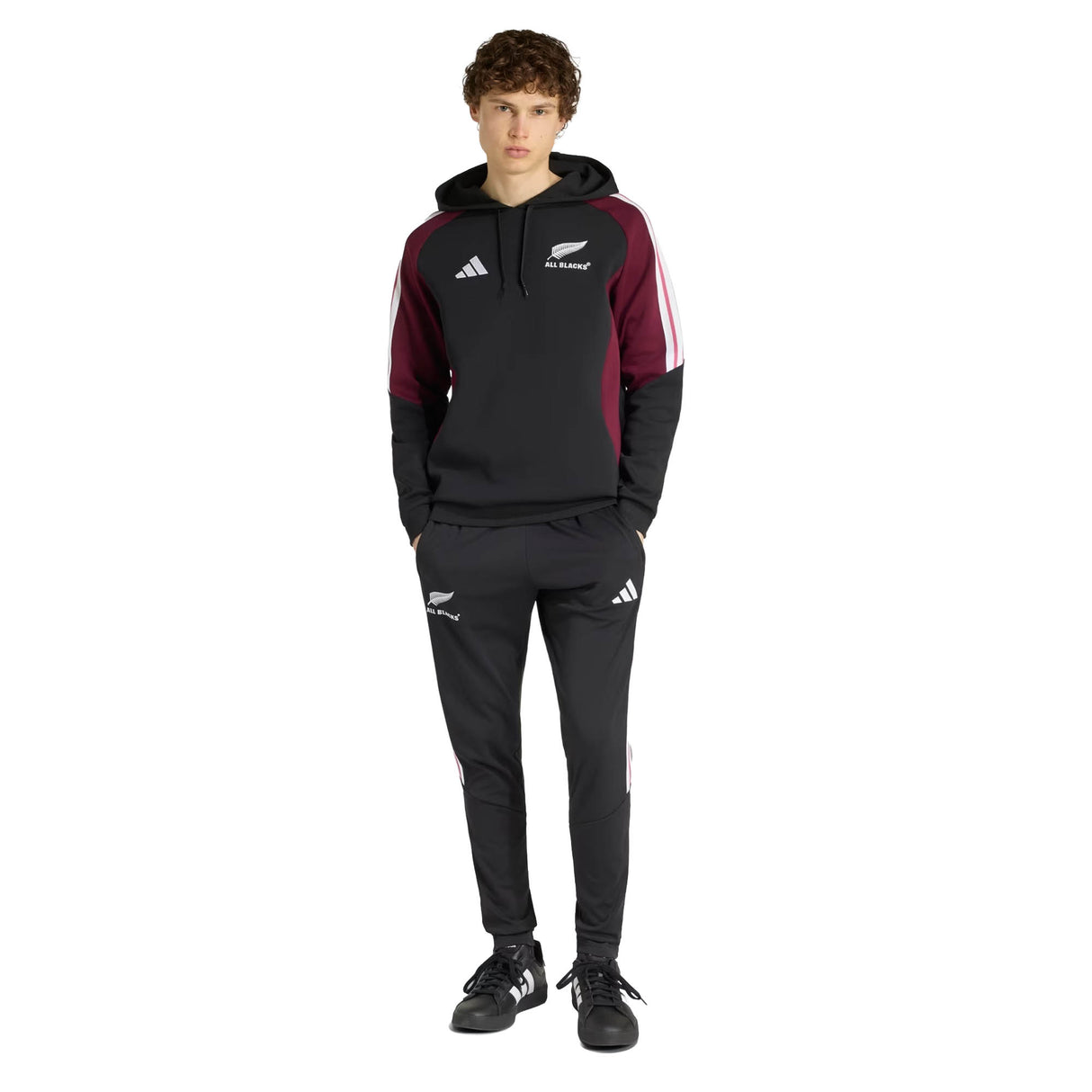 A person stands facing forward with hands in pockets, wearing the adidas All Blacks 27/28 Hoodie in black and maroon along with matching track pants, against a plain white background.