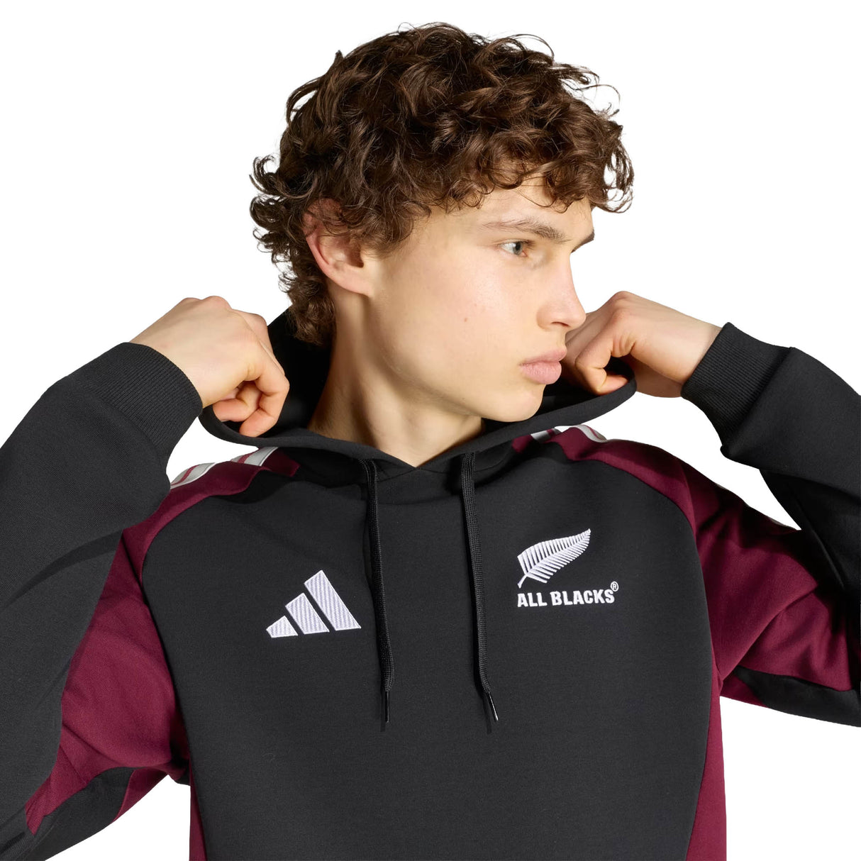 Wearing the All Blacks 27/28 Hoodie by adidas, a person adjusts the black hoodie with maroon sleeves featuring New Zealand rugby and adidas logos, looking to the side against a white background.