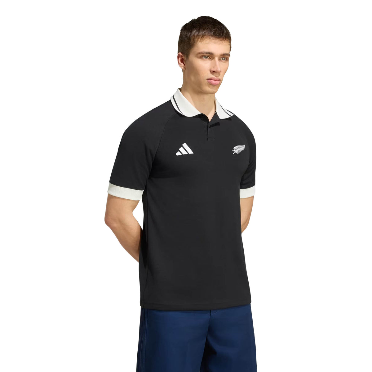 Wearing the All Blacks 26/27 Travel Polo by adidas—a black, short-sleeve collared shirt with white accents and two chest logos—a person stands with their hands behind their back against a plain background.