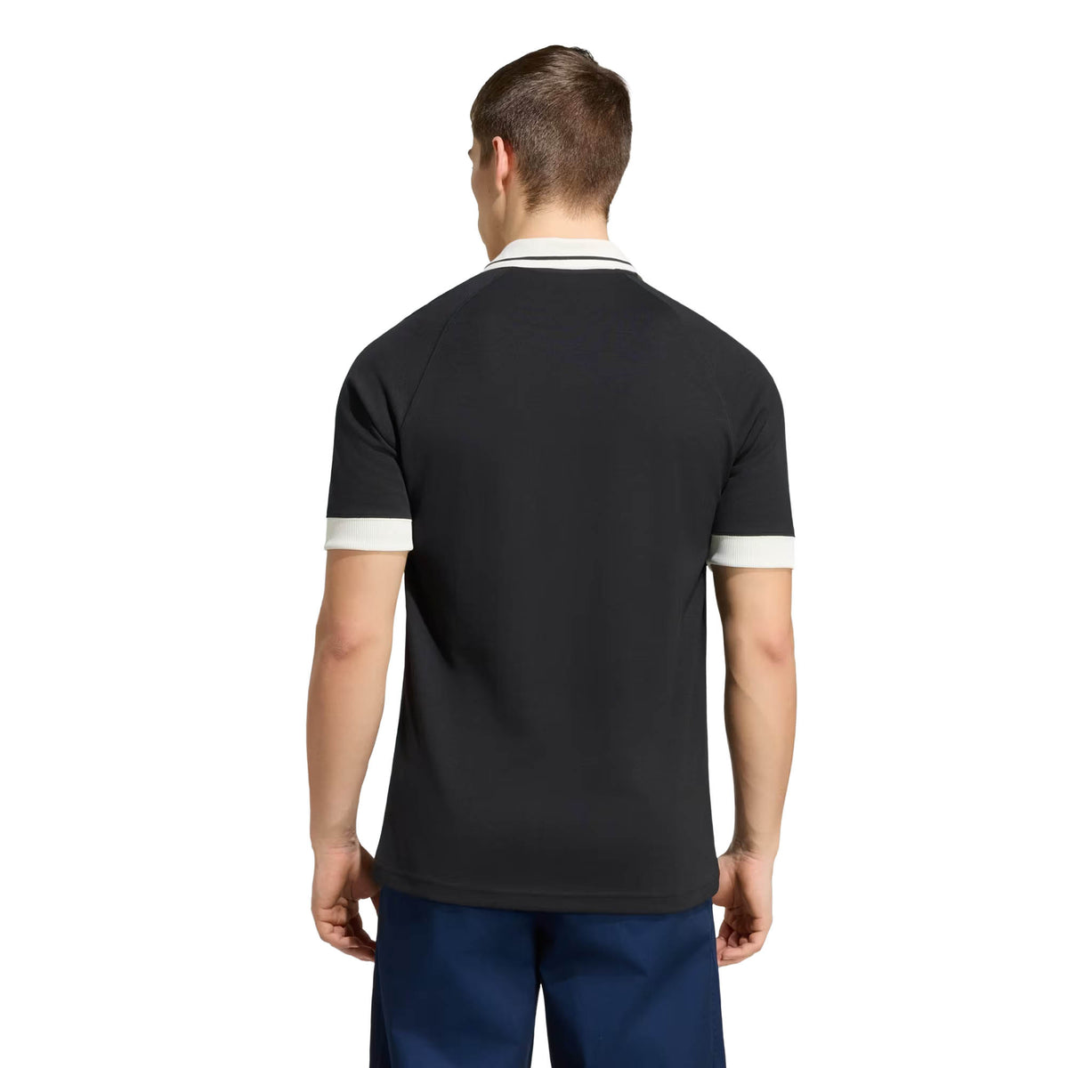 A man stands facing away, wearing the adidas All Blacks 26/27 Travel Polo with white trim on the collar and sleeves, paired with blue pants, against a plain white background.