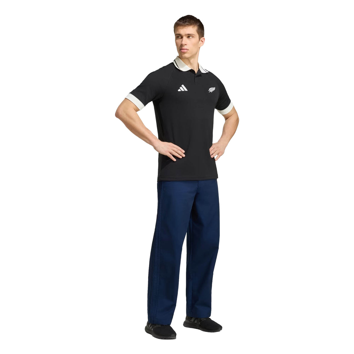 A man stands with hands on hips, wearing the adidas All Blacks 26/27 Travel Polo featuring team logos, along with blue pants and black shoes against a white background.