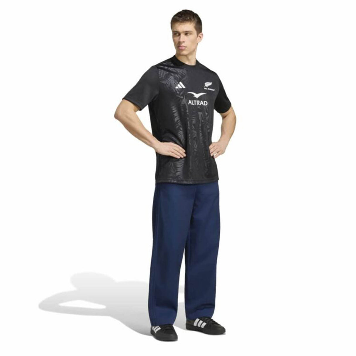 A person stands with hands on hips, wearing a black All Blacks Greatest Rivalry Supporters Tee by adidas—a tribute to New Zealand rugby—loose blue pants, and black adidas shoes, against a plain white background.