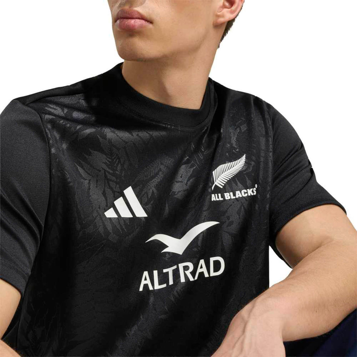 A person wearing the All Blacks Greatest Rivalry Supporters Tee by adidas, featuring adidas and Altrad logos along with the iconic fern emblem, shown from the shoulders up—a classic symbol of New Zealand rugby tradition.
