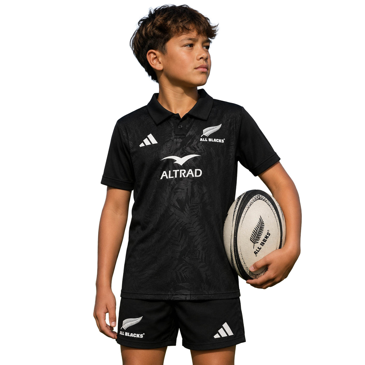 Clad in the Youth All Blacks Greatest Rivalry Replica Home Jersey by adidas, a young boy holds a rugby ball under his arm and stands against a plain white background—proudly representing New Zealand Rugby in this official adidas replica home jersey.