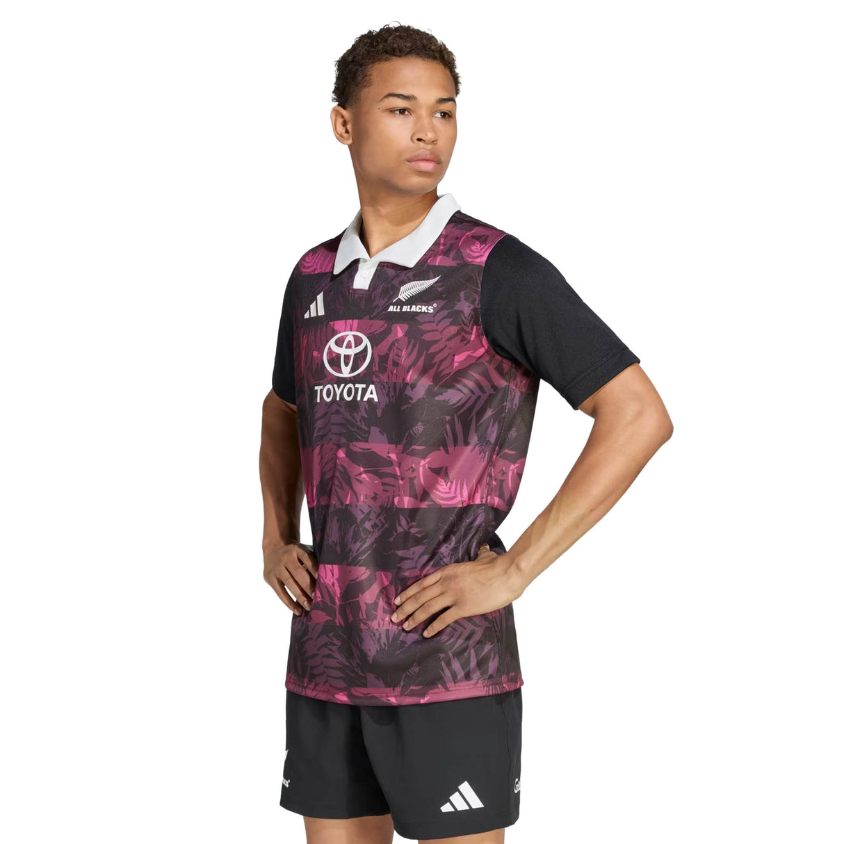 A person stands with hands on hips, wearing the All Blacks 26/27 Training Jersey – South Africa Tour by adidas, featuring a black and pink pattern with a white collar and adidas logos, paired with black shorts, against a plain white background.