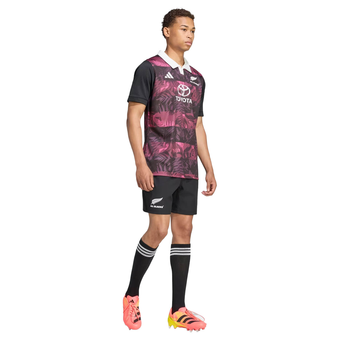 A person stands wearing the adidas All Blacks 26/27 Training Jersey – South Africa Tour, paired with black shorts, black knee-high socks with white stripes, and bright orange cleats against a white background—a look that echoes the official kit for the South Africa tour.