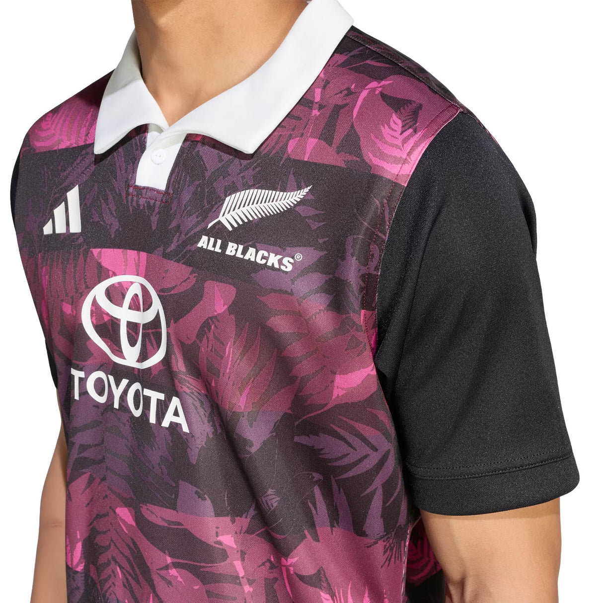 Wearing the All Blacks 26/27 Training Jersey - South Africa Tour by adidas, a person shows off a stylish look inspired by the team’s recent tour of South Africa, with pink and purple leaf patterns, a white collar, and the Toyota sponsor logo.