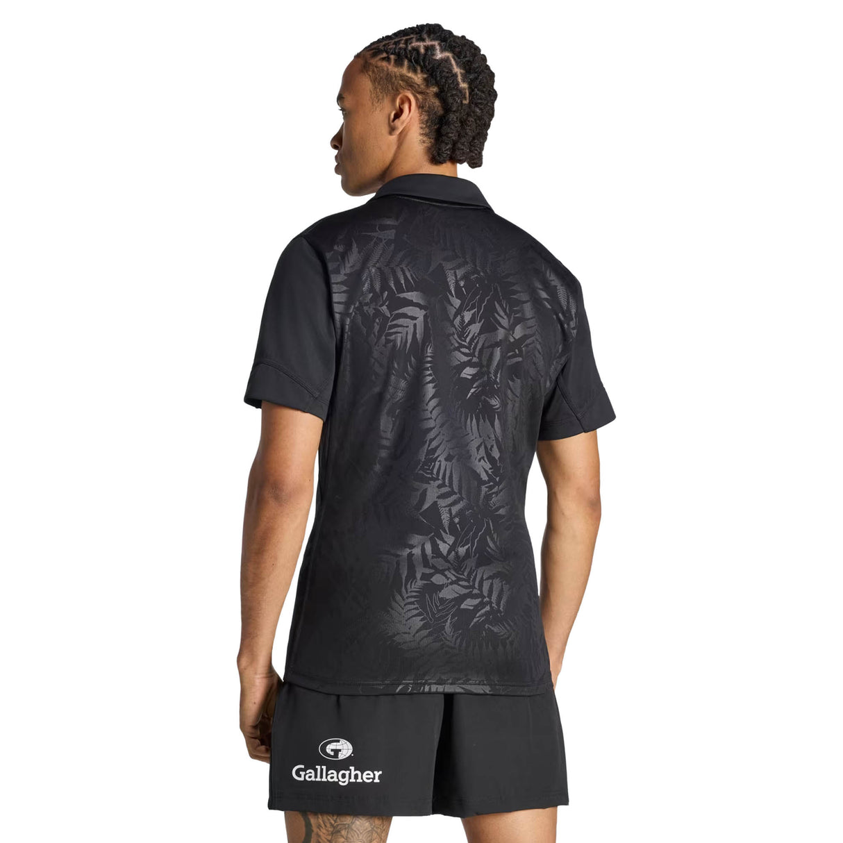 A person with braided hair stands with their back to the camera, wearing the All Blacks Greatest Rivalry Performance Home Jersey by adidas and matching black shorts featuring the Gallagher logo, evoking the iconic style of a classic All Blacks jersey.