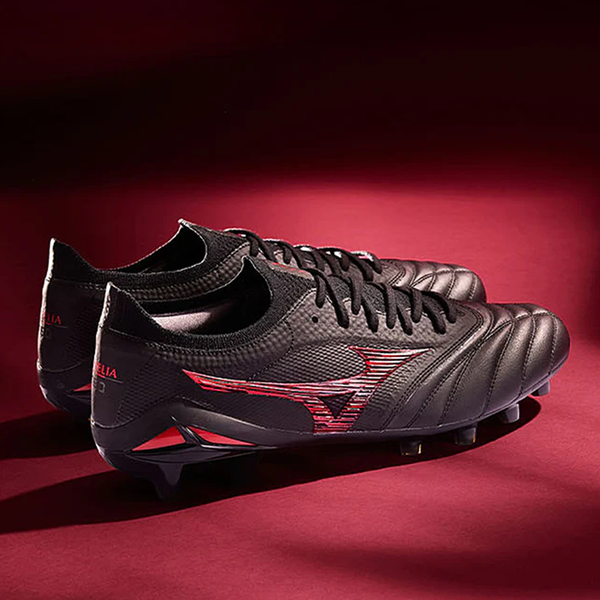 A pair of Mizuno Morelia Neo IV Beta FG Made in Japan soccer cleats in Black/Morelia 40th Anniversary Red is shown side by side on a maroon background.