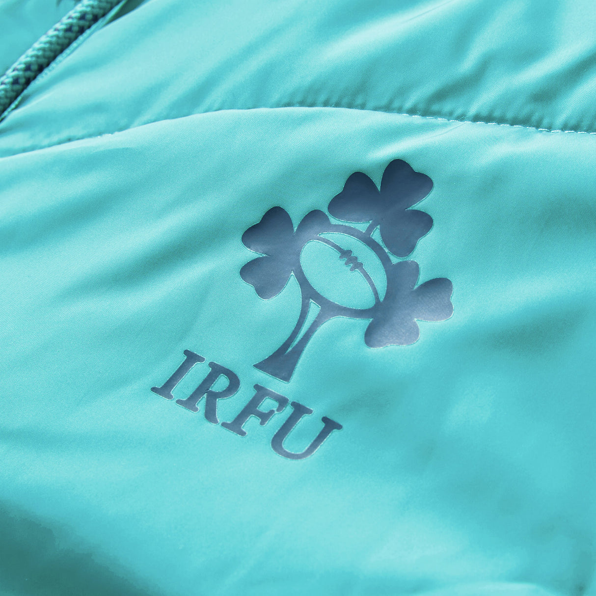 Close-up of a teal Ireland 26 Hybrid Padded Jacket by Canterbury, featuring the IRFU logo with a rugby ball and shamrock design printed above the letters “IRFU.”.