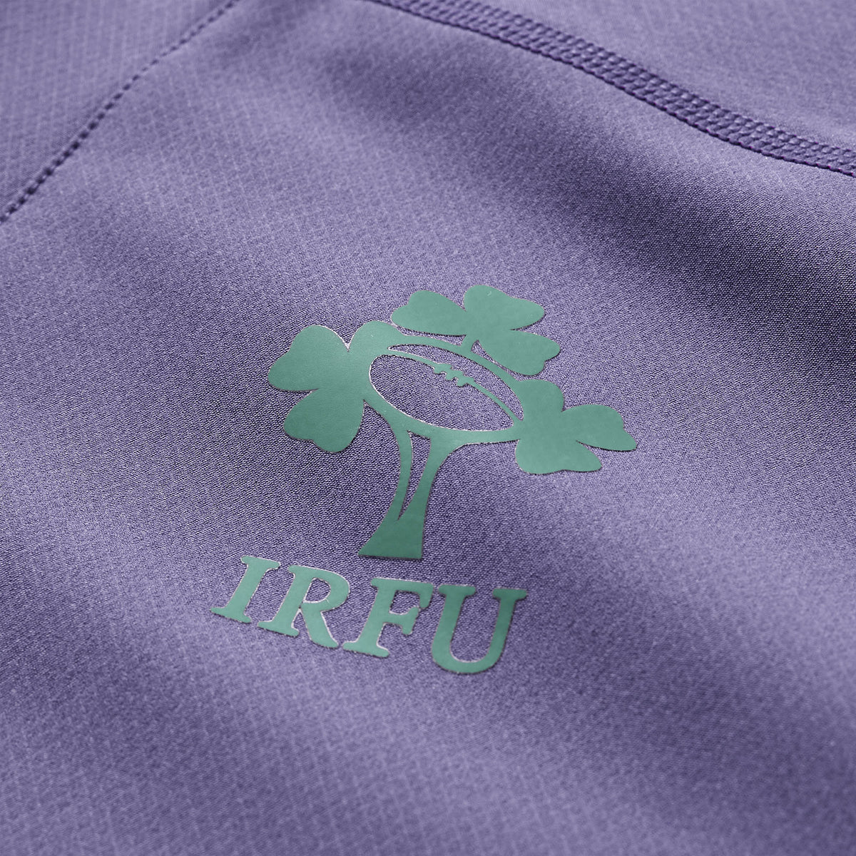Close-up of a green IRFU logo with a rugby ball and shamrock on textured purple fabric, showcasing the premium quality of the Ireland 26 Tech Drill Top by Canterbury from the official IRFU training wear collection.