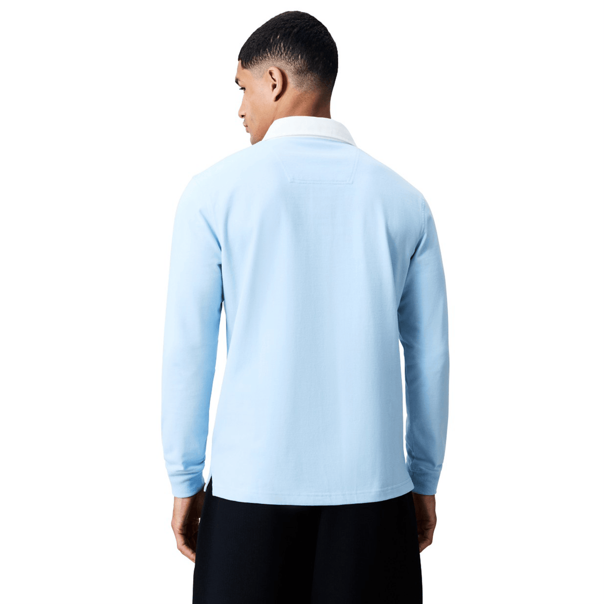 A man stands with his back to the camera wearing a Canterbury Solid Dye Rugby Jersey in light blue with a white collar and black pants, set against a plain white background.