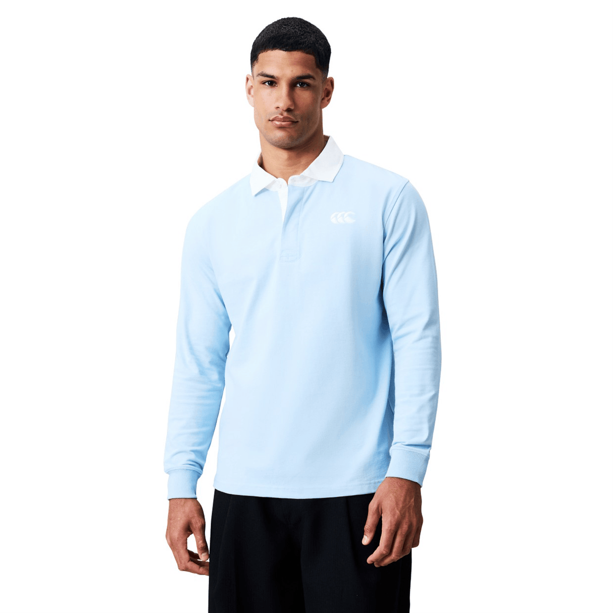 A man wearing the Canterbury Solid Dye Rugby Jersey by Canterbury, featuring a light blue long-sleeve design with a white collar and chest logo, paired with black pants, stands against a plain white background.