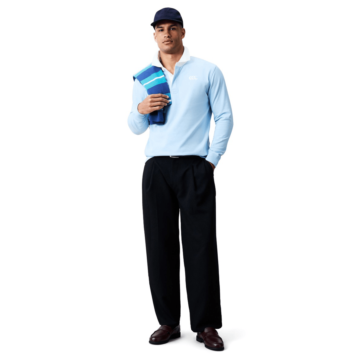 A man faces forward against a white background, wearing the Canterbury Solid Dye Rugby Jersey by Canterbury in blue, dark trousers, a navy cap, loafers, and holding a folded striped towel over his left shoulder.
