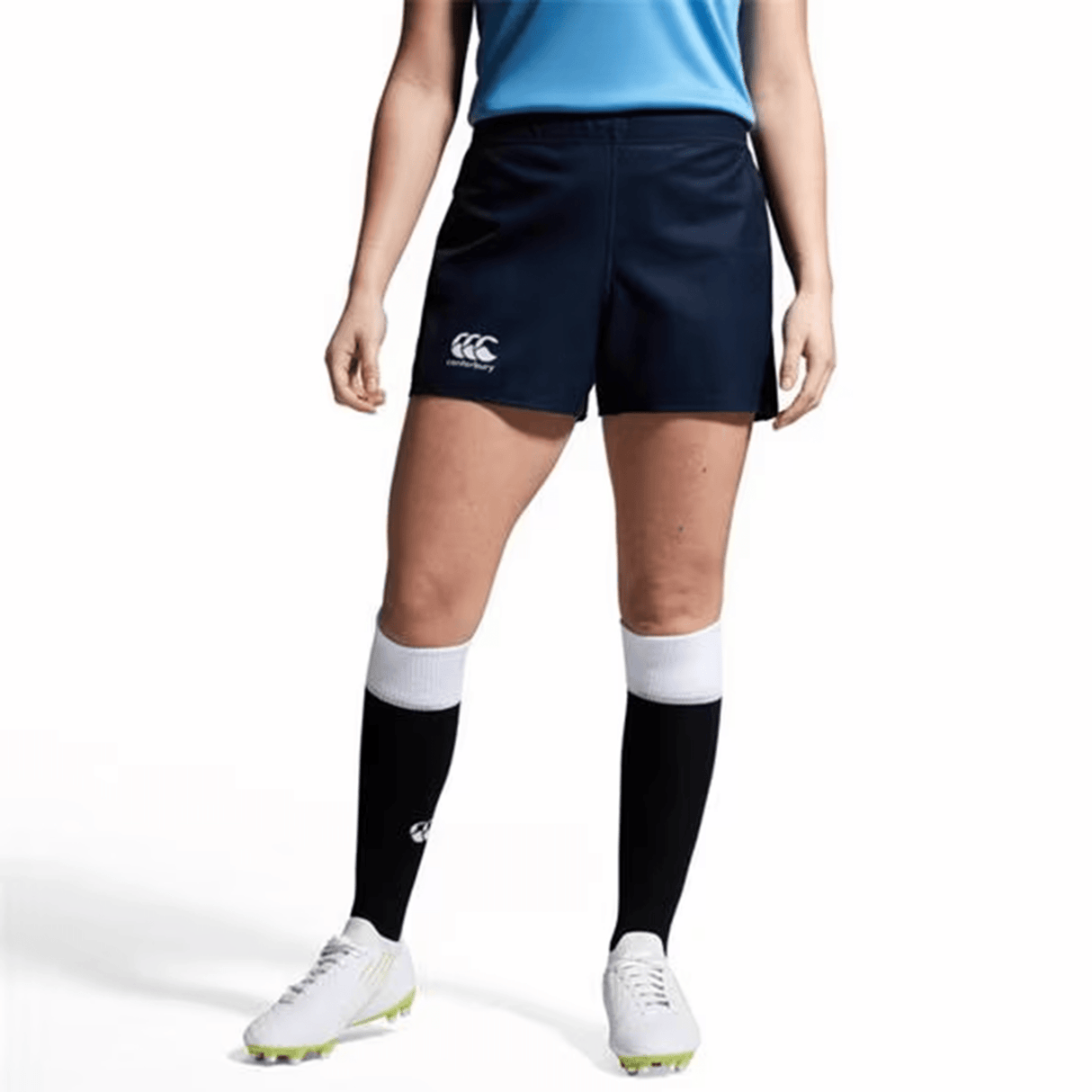 A person wearing a blue shirt with the Canterbury logo, Women's Canterbury Yokohama Shorts, white cleats, and black knee-high socks stands against a plain background.