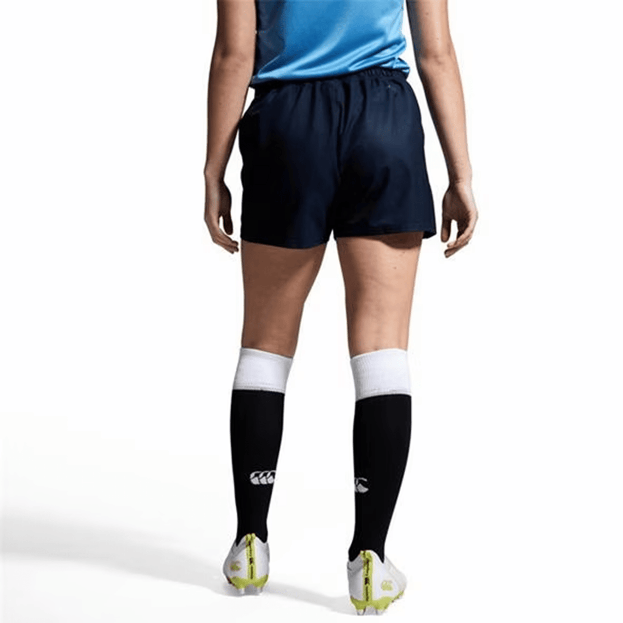 A person in a blue Vapodri fabric shirt, Canterbury's black Yokohama Shorts, knee-high black and white socks, and neon green-trimmed cleats with a subtle Canterbury logo stands on a white background.