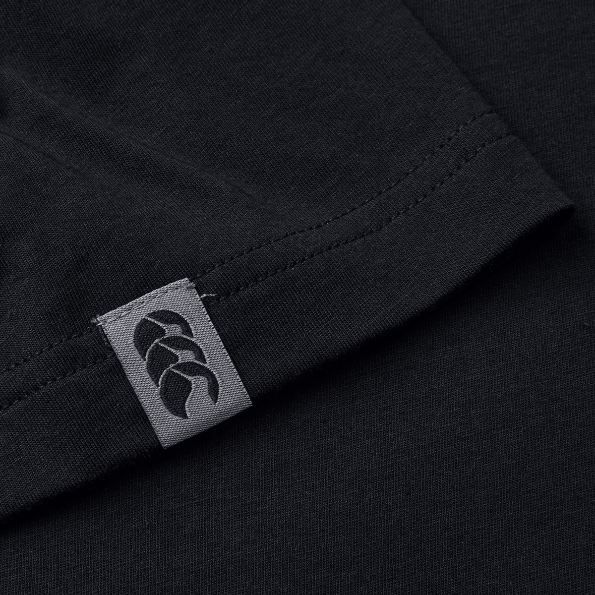 A close-up of the Canterbury Force T-Shirt shows its black fabric with a gray tag and braided hem design, celebrated for its relaxed fit and versatility.