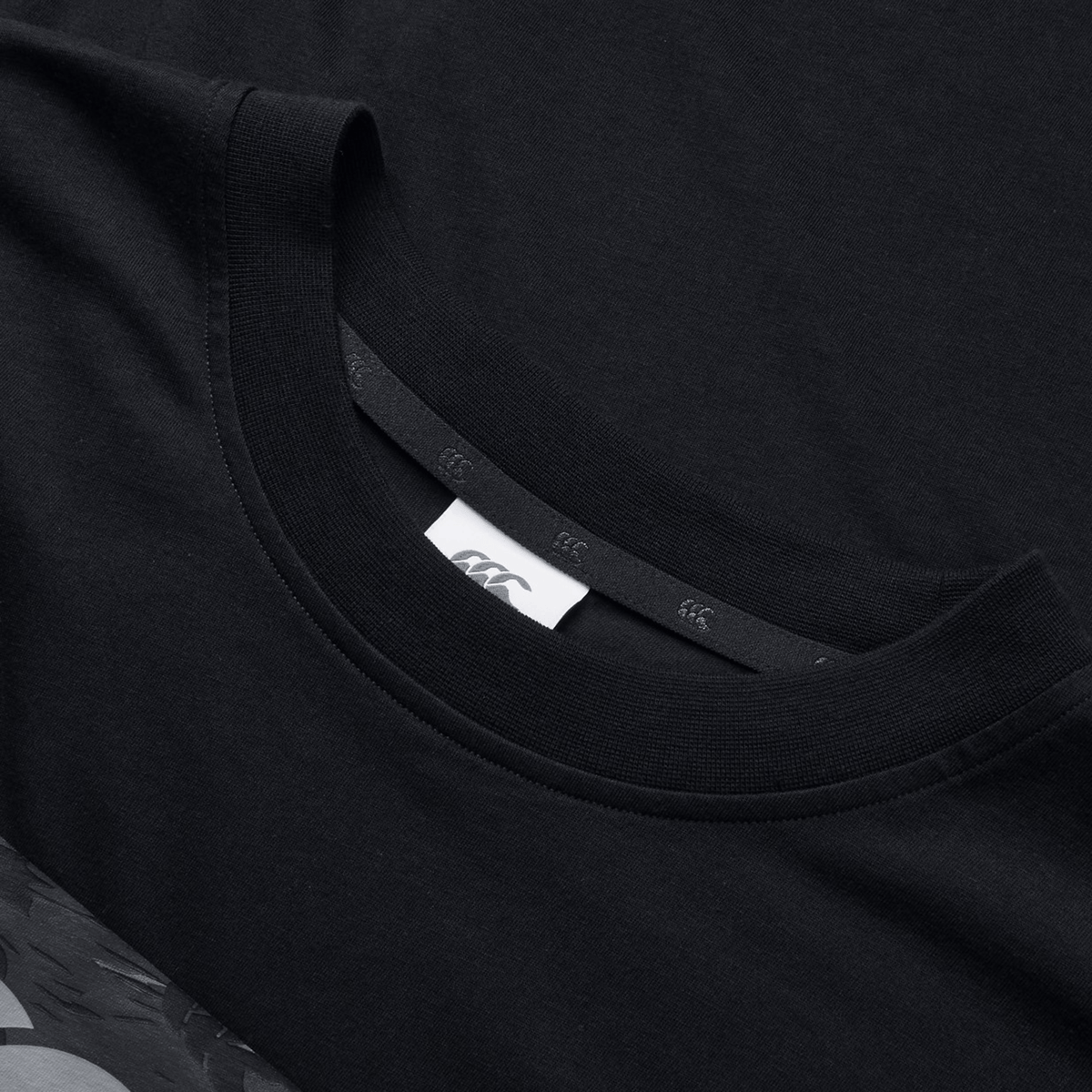 Close-up of the Canterbury Force T-Shirt in black, featuring a prominent neck tag with multiple "A" logos. Its relaxed fit seamlessly blends with the matching fabric background, underscoring its versatility for any wardrobe inclusion.