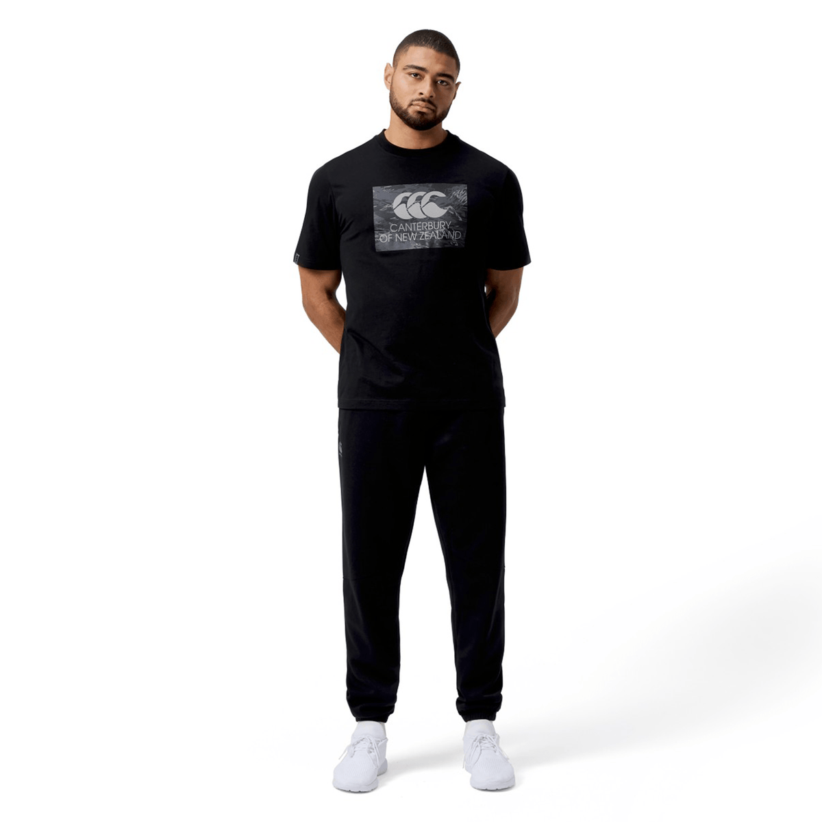 A man stands confidently in a relaxed fit Canterbury T-Shirt, hands casually behind his back against a crisp white background, effortlessly showcasing its versatility.