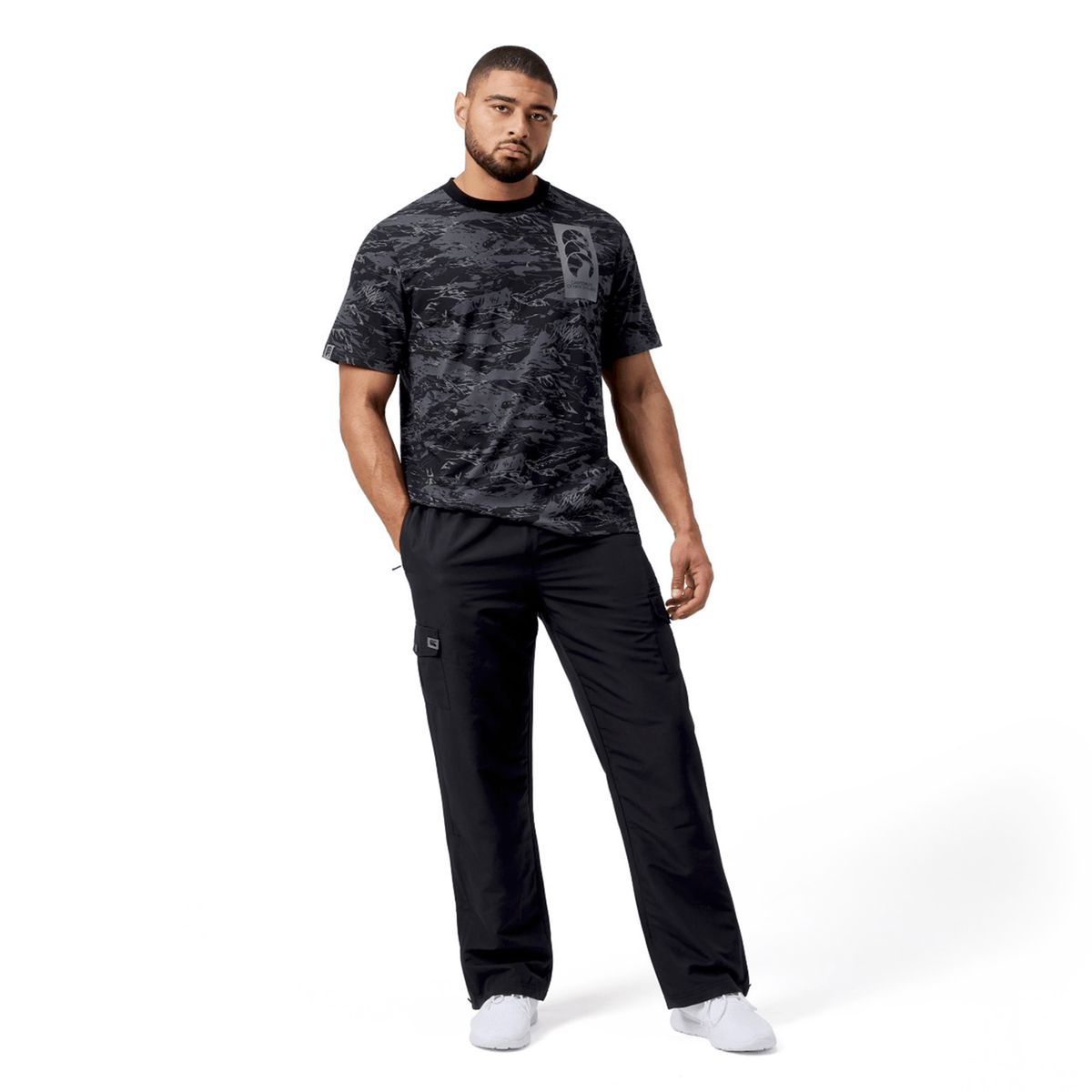 A man in a Canterbury Force AOP T-Shirt, black pants, and white sneakers stands with one hand in his pocket against a plain white background.