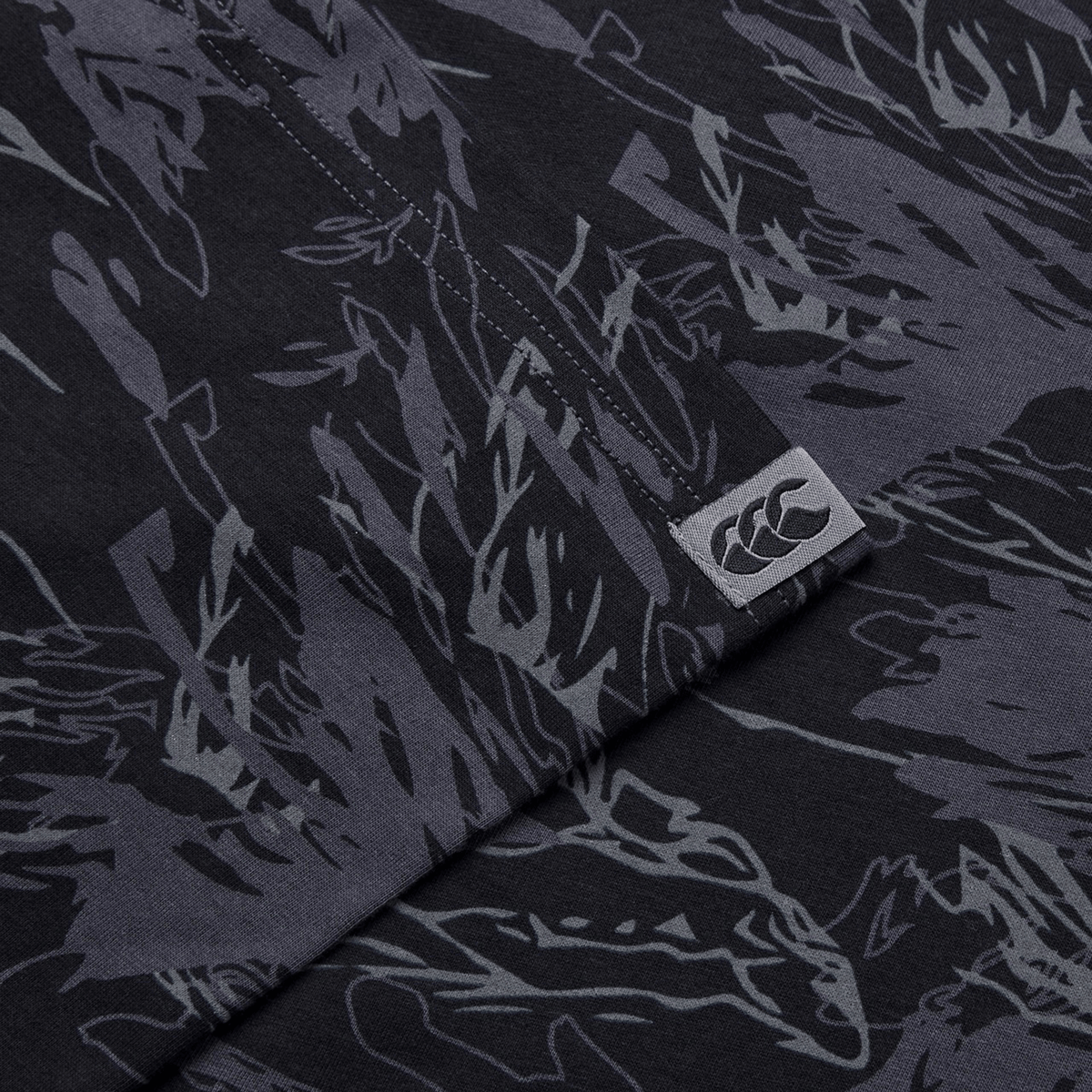 Close-up of a dark fabric with Canterbury's Force AOP T-Shirt camouflage print, featuring a small logo with overlapping "C" shapes in white on a rectangular tag.