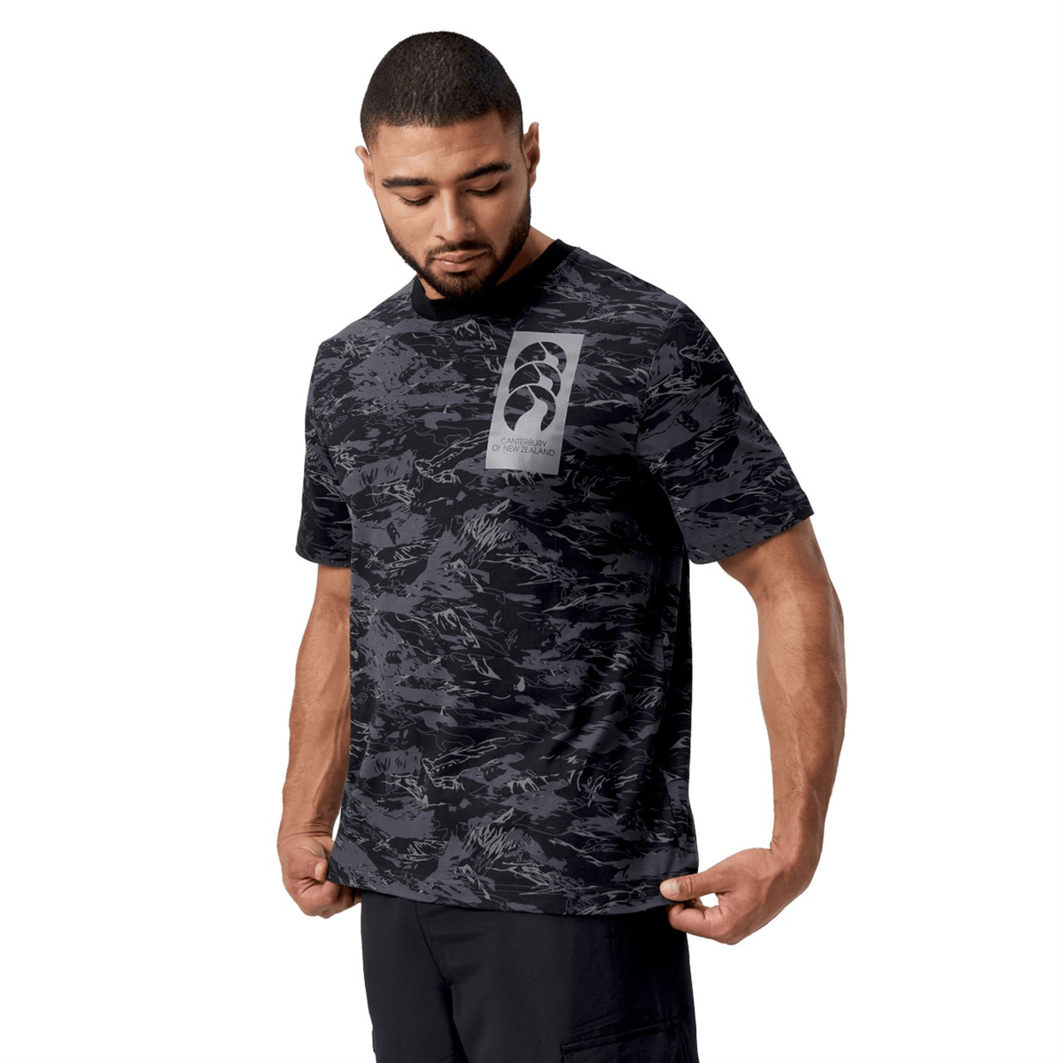 A man in a Canterbury Force AOP T-Shirt, with a CCC camouflage print and modern crew neck, examines the hem while standing against a plain white background.