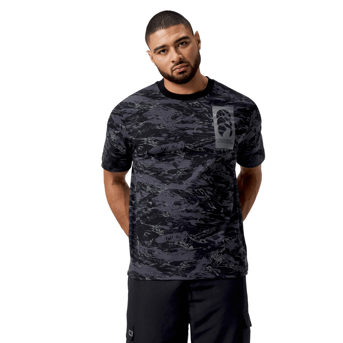 A man stands against a white background wearing the Canterbury Force AOP T-Shirt, featuring a black and gray CCC camouflage print and modern crew neck design, with a sleek chest logo.