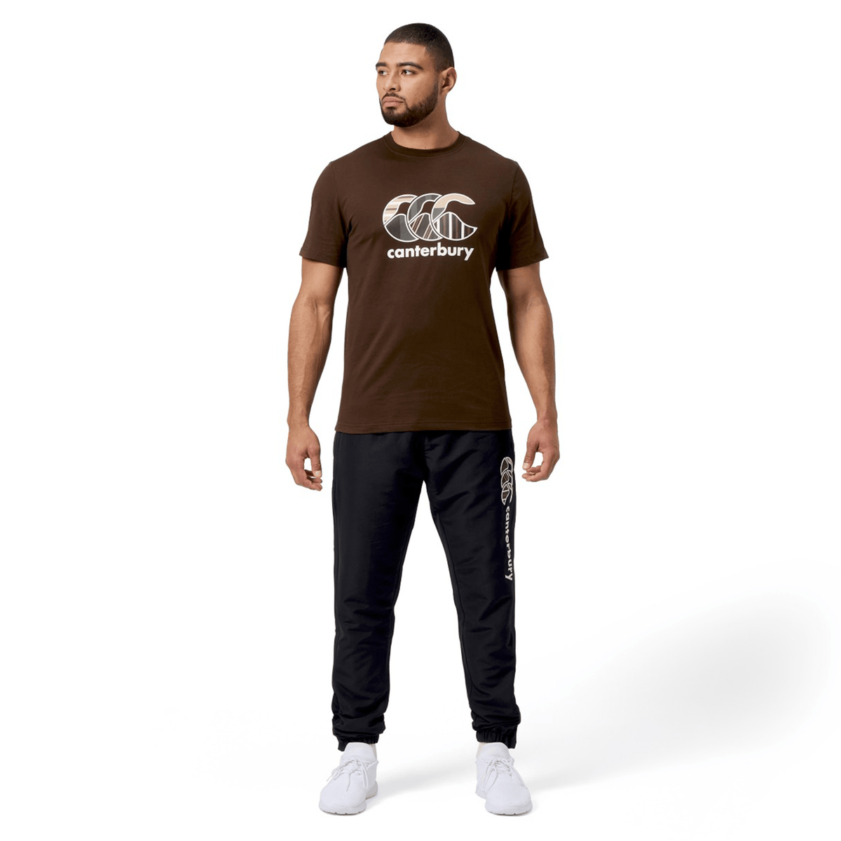 A man wears a vibrant Canterbury Uglies T-Shirt by Canterbury and black pants, facing slightly left with his hands at his sides against a white background.