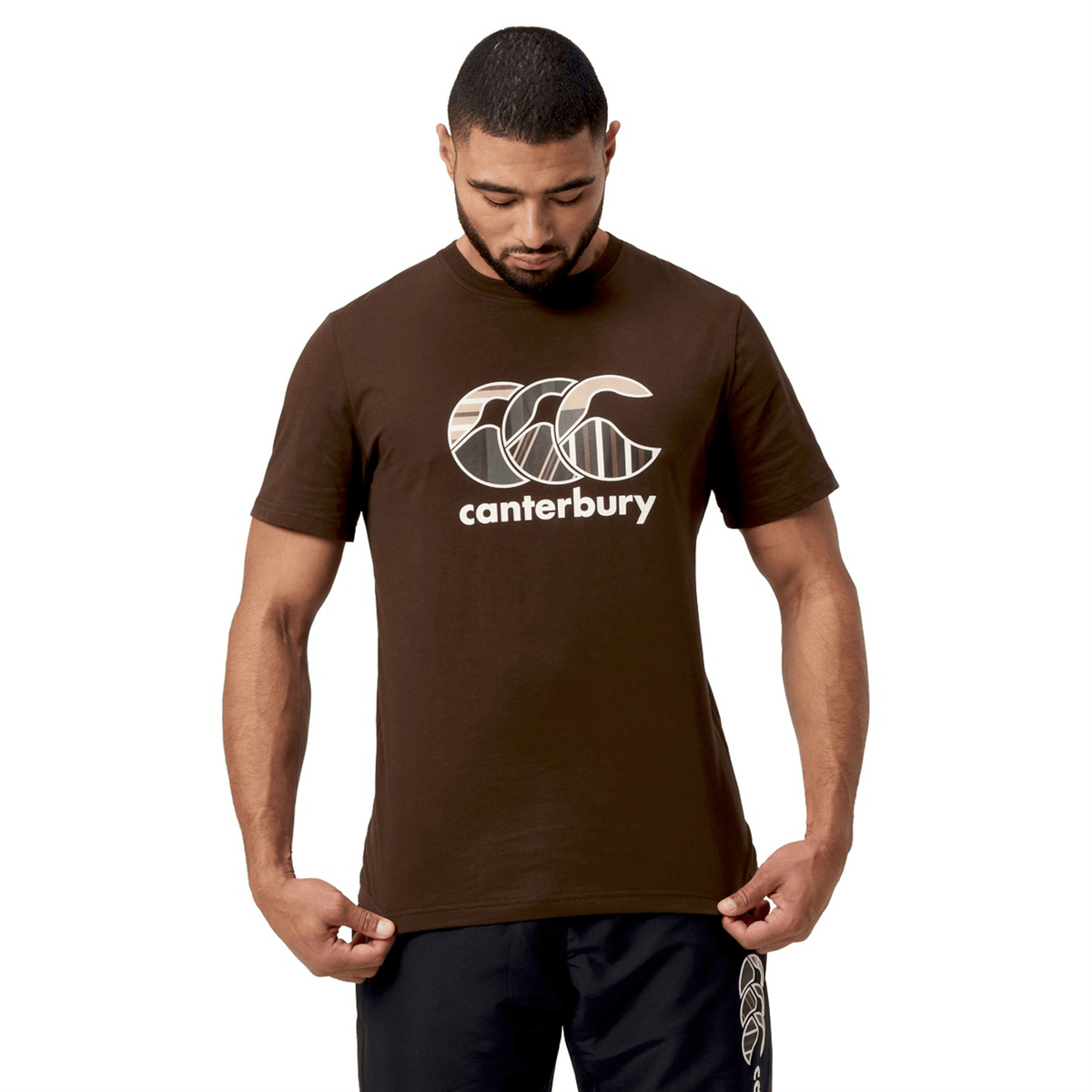 A man looking down wearing a brown Canterbury Uglies T-Shirt in premium cotton with a logo, along with black pants, set against a white background.