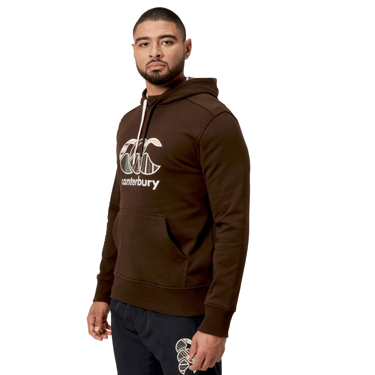 A person wearing a Canterbury Uglies Hoodie, featuring a brown color and a stylized CCC logo, stands slightly turned with hands by their sides and neutral expression.