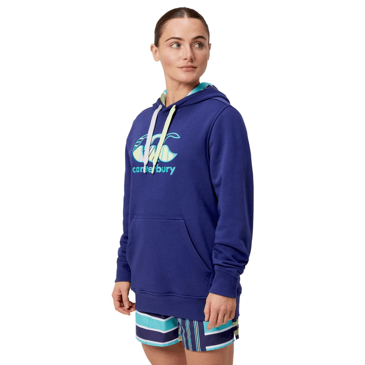 A person in a Canterbury Women's Uglies Hoodie with logo and striped shorts, standing against a white background, embodies Canterbury flair.