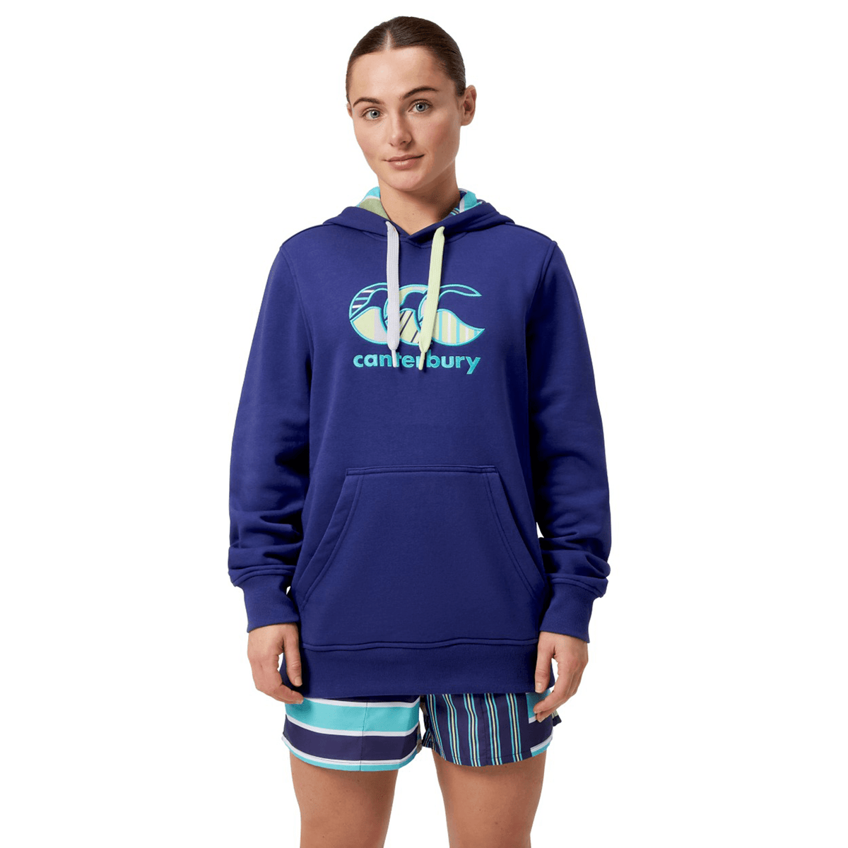 A person wears a Canterbury Women's Uglies Hoodie against a white background, styled with striped shorts that enhance their sporty look.