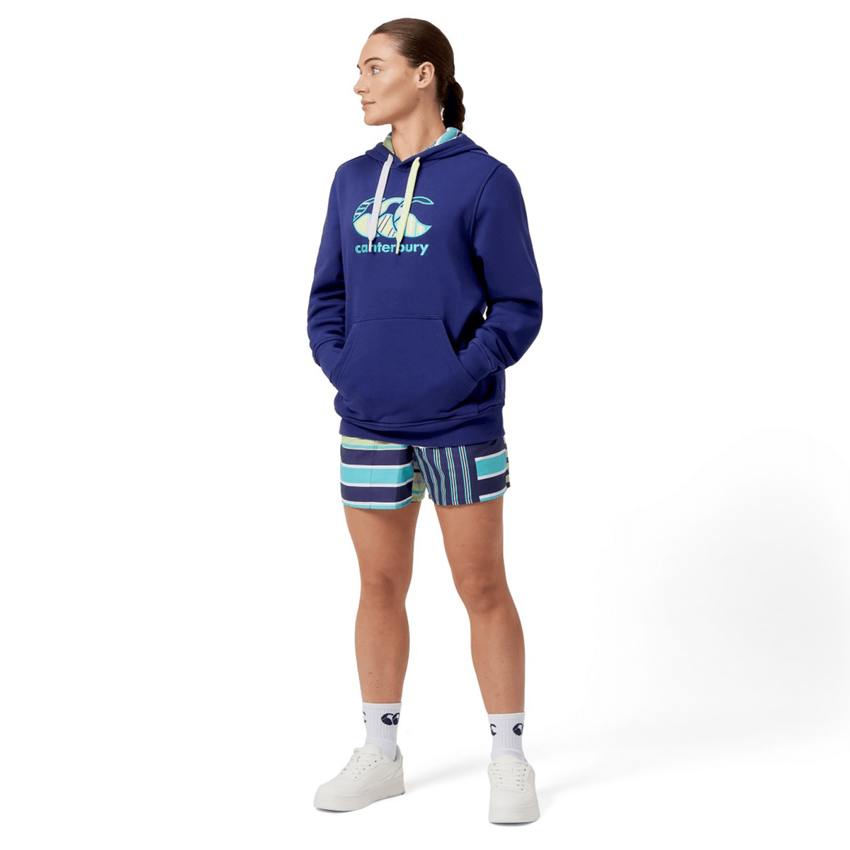 A person in a Canterbury Women's Uglies Hoodie in blue and striped shorts stands with hands in pockets, looking to the side. They're wearing white high-top sneakers and socks, exuding a laid-back vibe.
