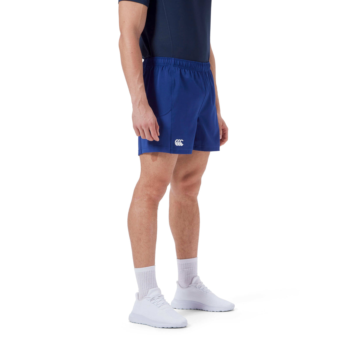 A person is wearing a black shirt, blue Canterbury Advantage Short 2.0 with a logo, white socks, and white shoes made from recycled content, standing against a plain white background.
