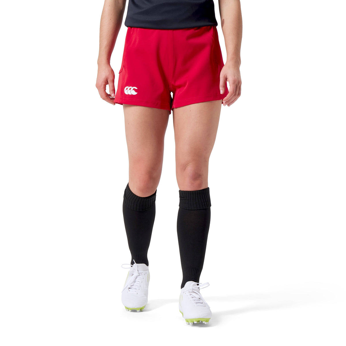 A person wearing Canterbury's Women's Advantage Short 2.0 in red, paired with black socks and white shoes, stands against a white backdrop. The outfit highlights performance quality and reflects a commitment to sustainability through its recycled content design.
