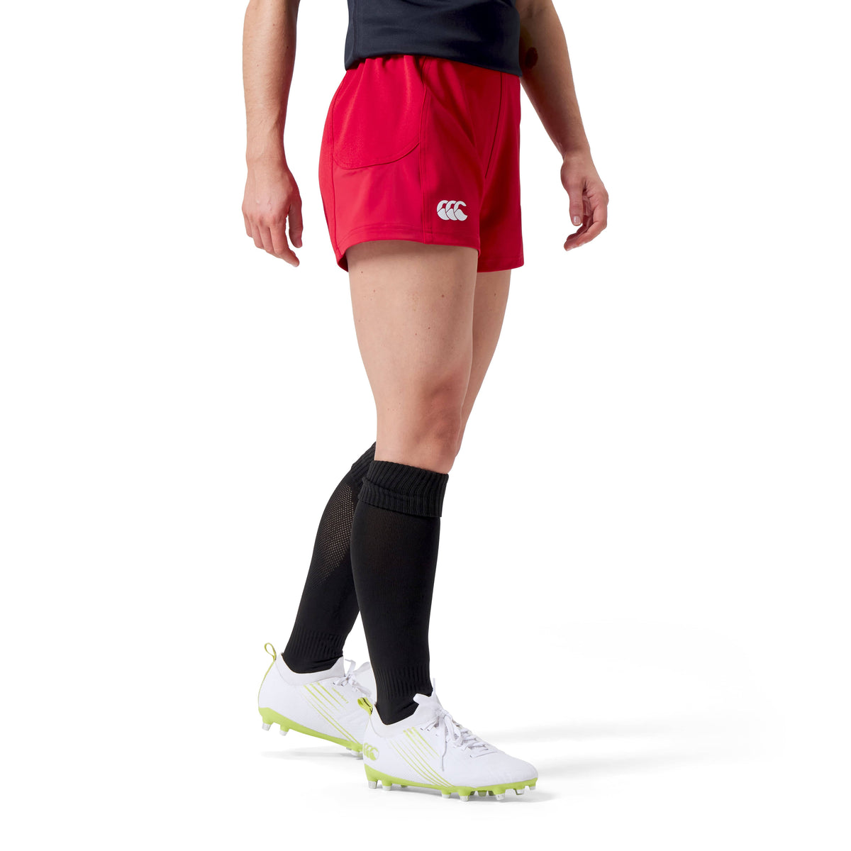 A woman in Canterbury Women's Advantage Short 2.0, black socks, white cleats, and a dark top stands on a white background, showcasing performance and sustainability.