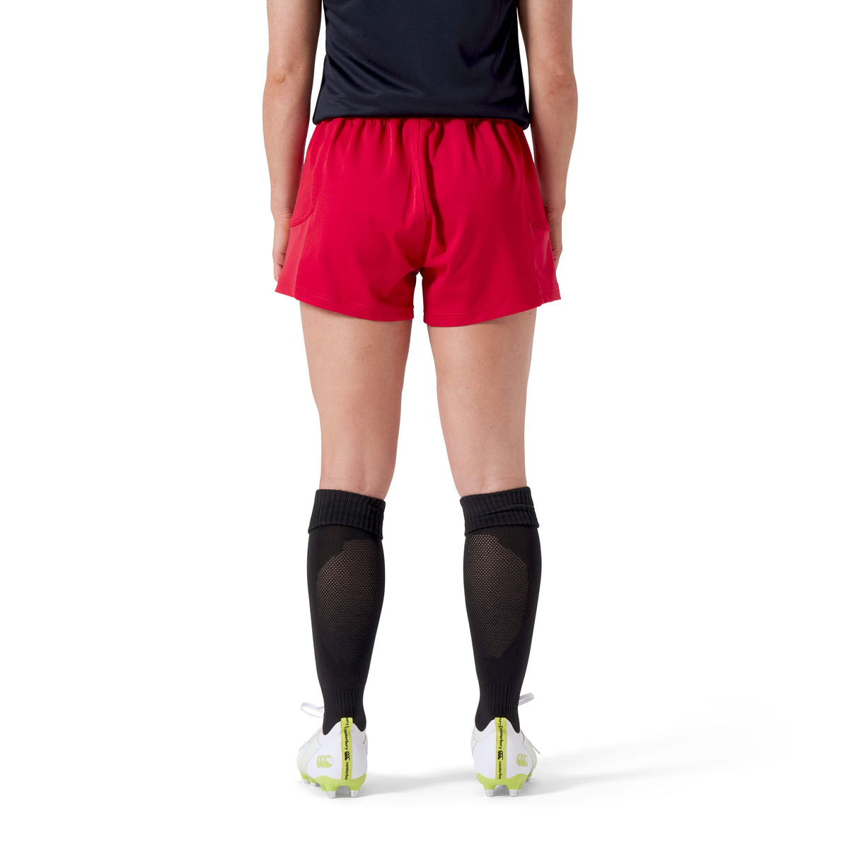 A person stands with their back to the camera, embodying performance in a black shirt, Canterbury's Women's Advantage Short 2.0 in red, black socks, and white cleats made with recycled content.