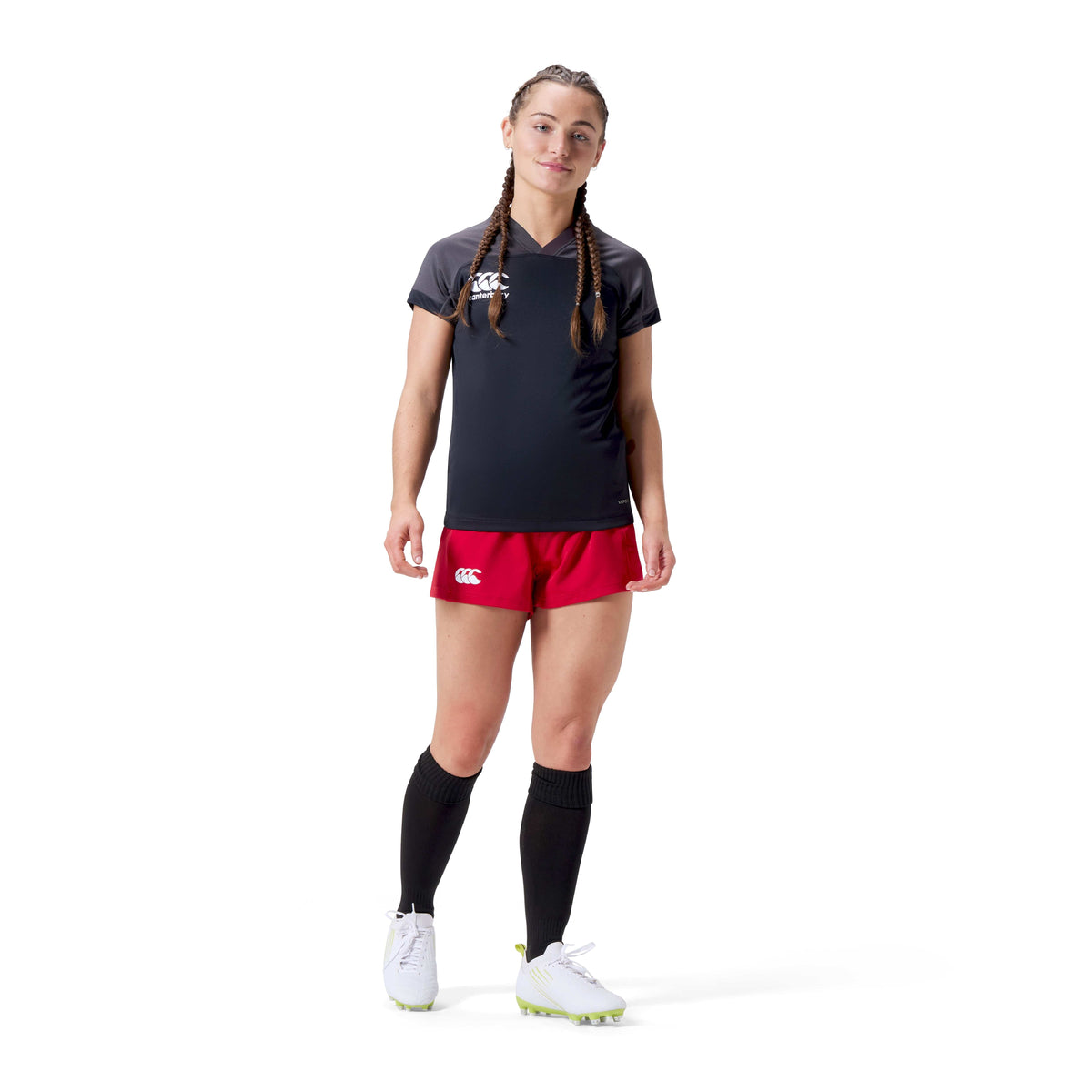 A woman in a black sports shirt, Canterbury's Women's Advantage Short 2.0 in red, knee-high black socks, and white cleats stands against a white background. Her hair is styled in two braids. The outfit blends performance with sustainability using recycled materials for an eco-friendly fashion statement.