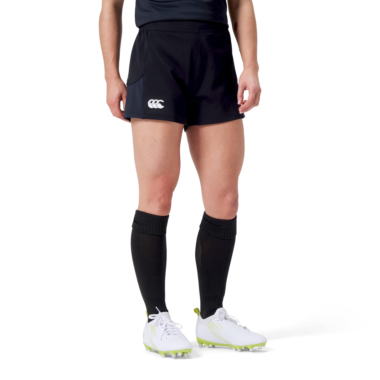 A person in a black sports uniform made from recycled materials, wearing Women's Canterbury Advantage Short 2.0, long socks, and white cleats, stands against a plain background.