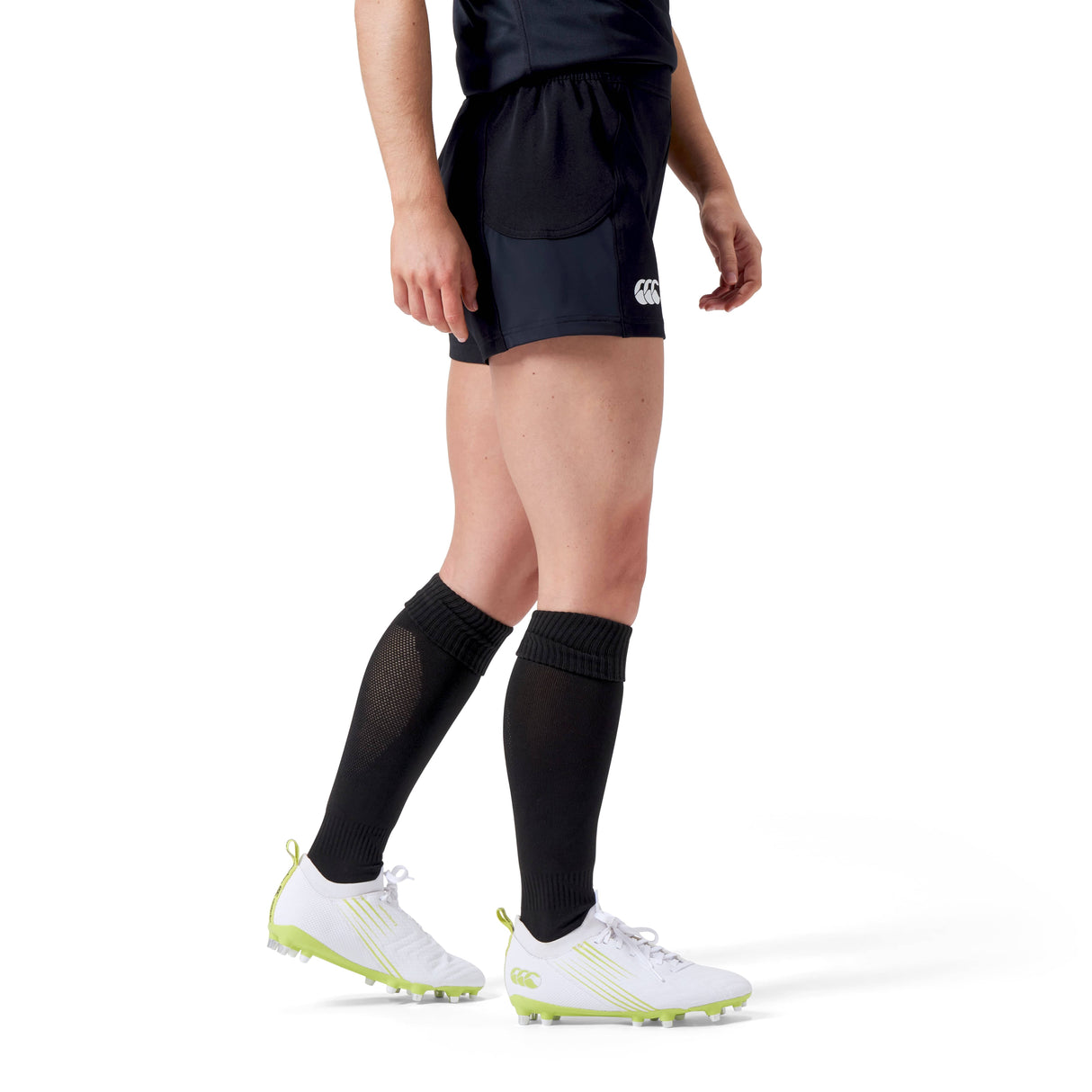A woman strides confidently in Canterbury Women's Advantage Short 2.0, long black socks, and white cleats with light green soles, showcasing a commitment to performance and sustainability through the use of recycled content.