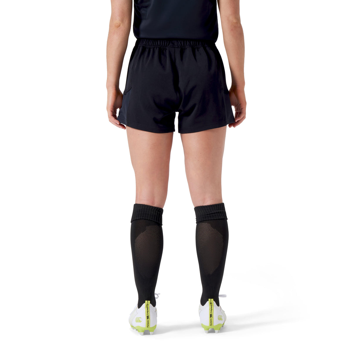 A person in Canterbury Women's Advantage Short 2.0—a black sports outfit made from recycled materials—stands on a white background, sporting knee-high socks and white cleats, showcasing performance and sustainability.