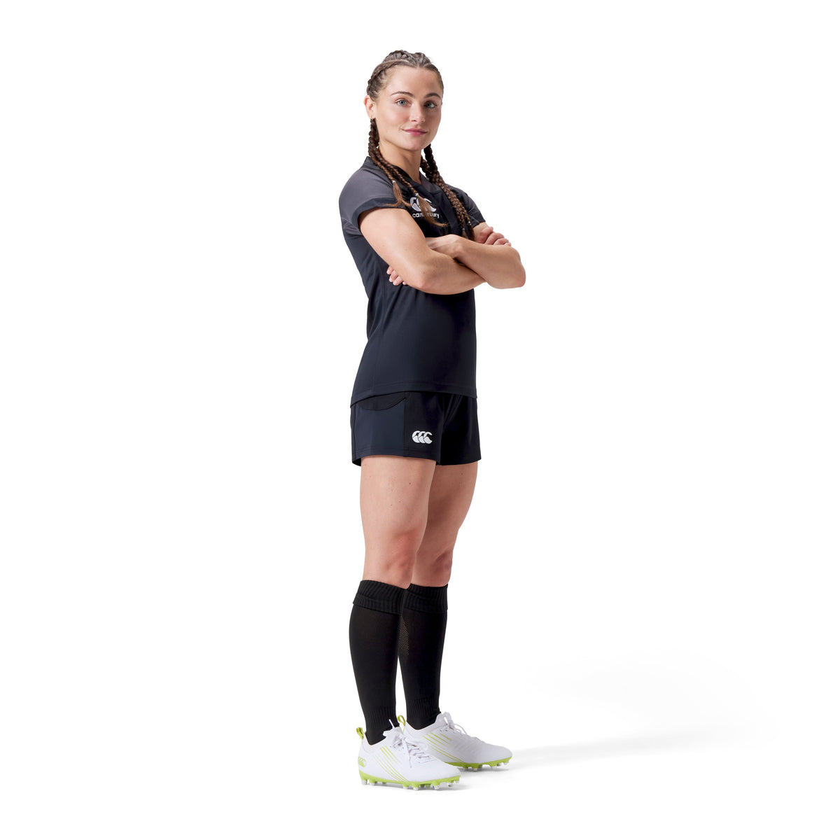 A person with braided hair stands confidently, arms crossed, wearing Canterbury's Women's Advantage Short 2.0 in black, engineered for performance and sustainability, along with long socks and white cleats.