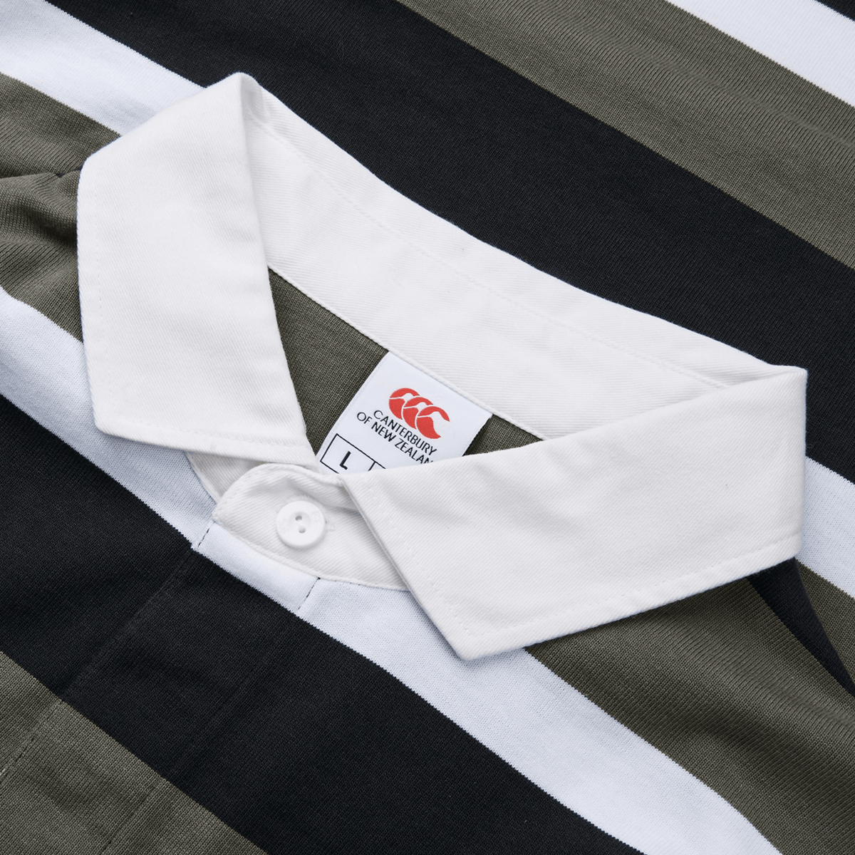 Close-up of the Canterbury Yarn Dye Long Sleeve Striped Rugby Jersey, showcasing its crisp white collar and black, olive green, and white stripes. A visible tag reveals the brand (Canterbury) and size details.